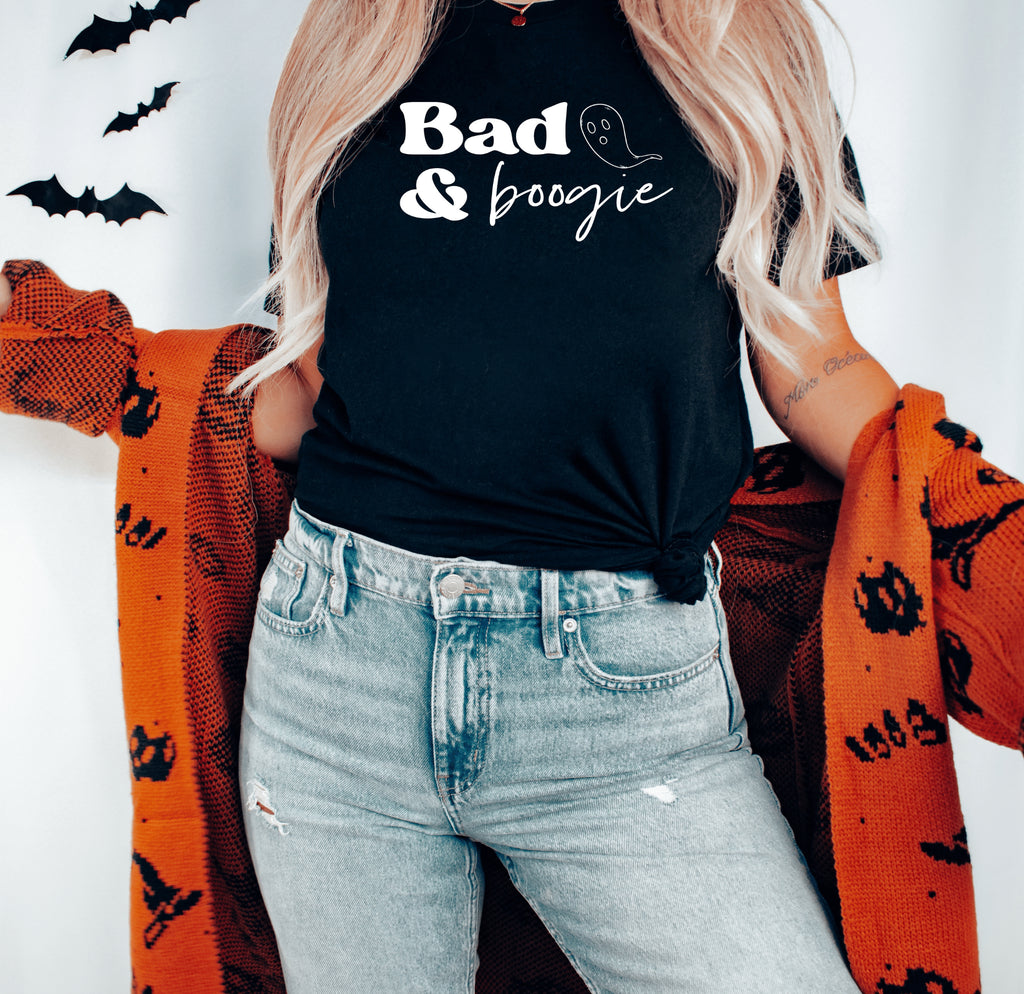 adult woman with blonde hair, wearing a black halloween t-shirt with white text and a ghoast with orange ghost pictures on the cardigan with light wash blue jeans with bats in the bacground, mock photo