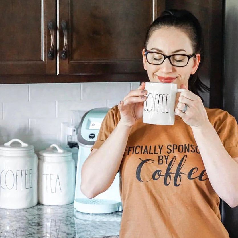 woman smiling holding a rae dunn coffee mug smelling the coffee in officially sponsored by coffee shirt screen print design