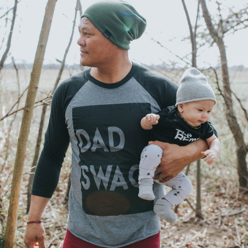 dad in grey and blag raglan shirt matching in a beanie hat with baby and baby wearing remix onesie, dad holding baby with one arm looking like a super hero