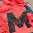 flat lay coral hoodie with black ink that says mother with a varsity letter M laying on top of skinny blue jeans for a cute cozy outfit for mothers