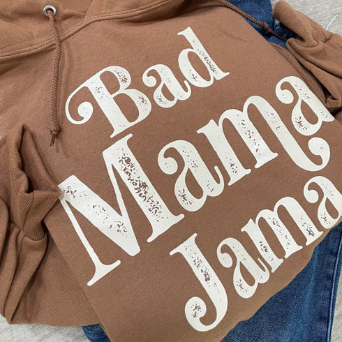 close up of a flat lay of a brown hoodie with cream ink screen print design that says bad mama jama, laying on some cute everyday blue jeans for a cozy outfit for moms with a cute saying. hoodie color also called pecan