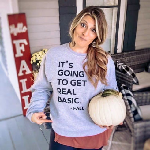 woman wearing grey sweatshirt with black text with the design its going to get real basic fall, holding a pumpkin and hell fall decorations on her porch