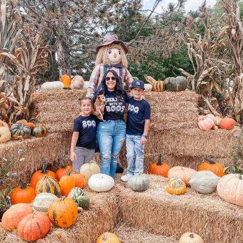 halloween theme shirts worn by mom and kids at a fall festival with fall decorations of hay stacks and pumpkins and a scare crow showing the perfect fall activity theme for family photo and matching shirts that says rollin with my boos and the kids wearing boos
