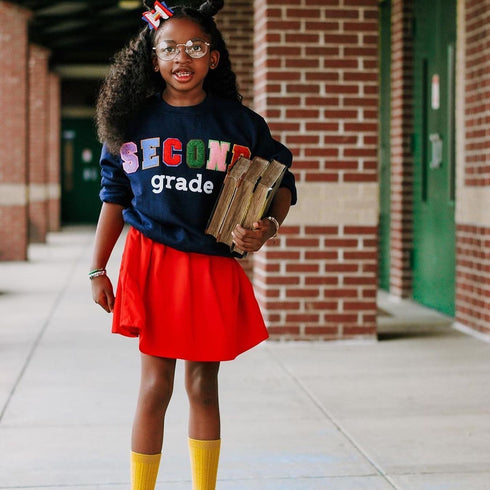 little girl in navy custom sweatshirt with chenille patches that are multi colored in a red skirt, black girl magic representing education holding books in yellow socks