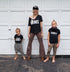mom and two boys in front of a garage wearing matching shirts with screen print design that says moms shorty and shorter shorty and mom wearing the original hat with a long elsa braid